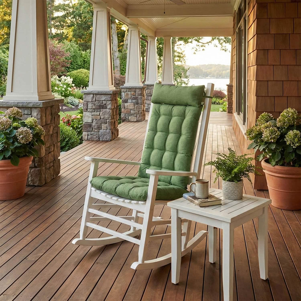 Rocking chair with green cushions on a wooden porch with potted plants and a view of a lake.