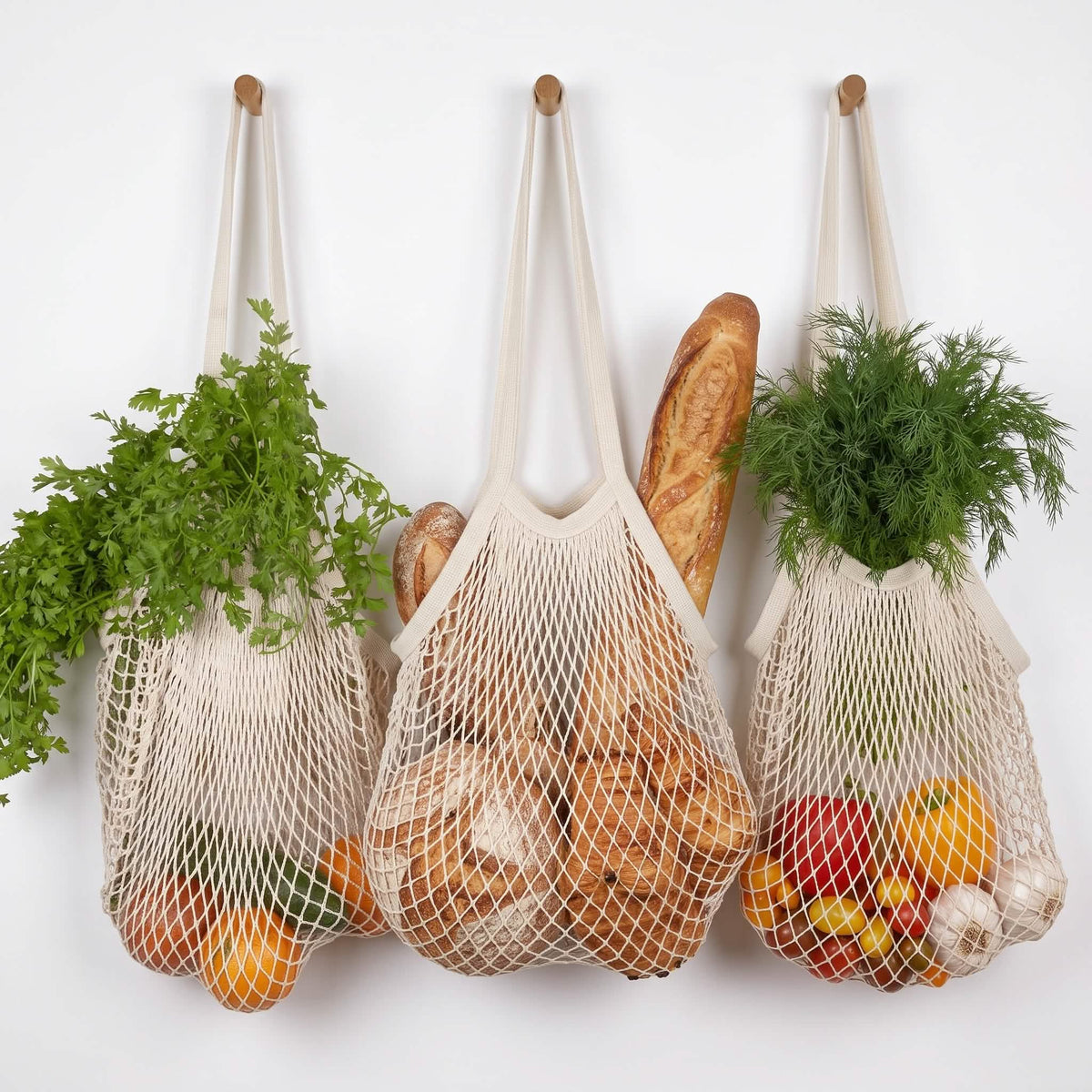 Three french market grocery bags of cotton mesh hanging from wooden hooks on a white wall, filled with fresh greens, bread, and colorful fruits and vegetables.