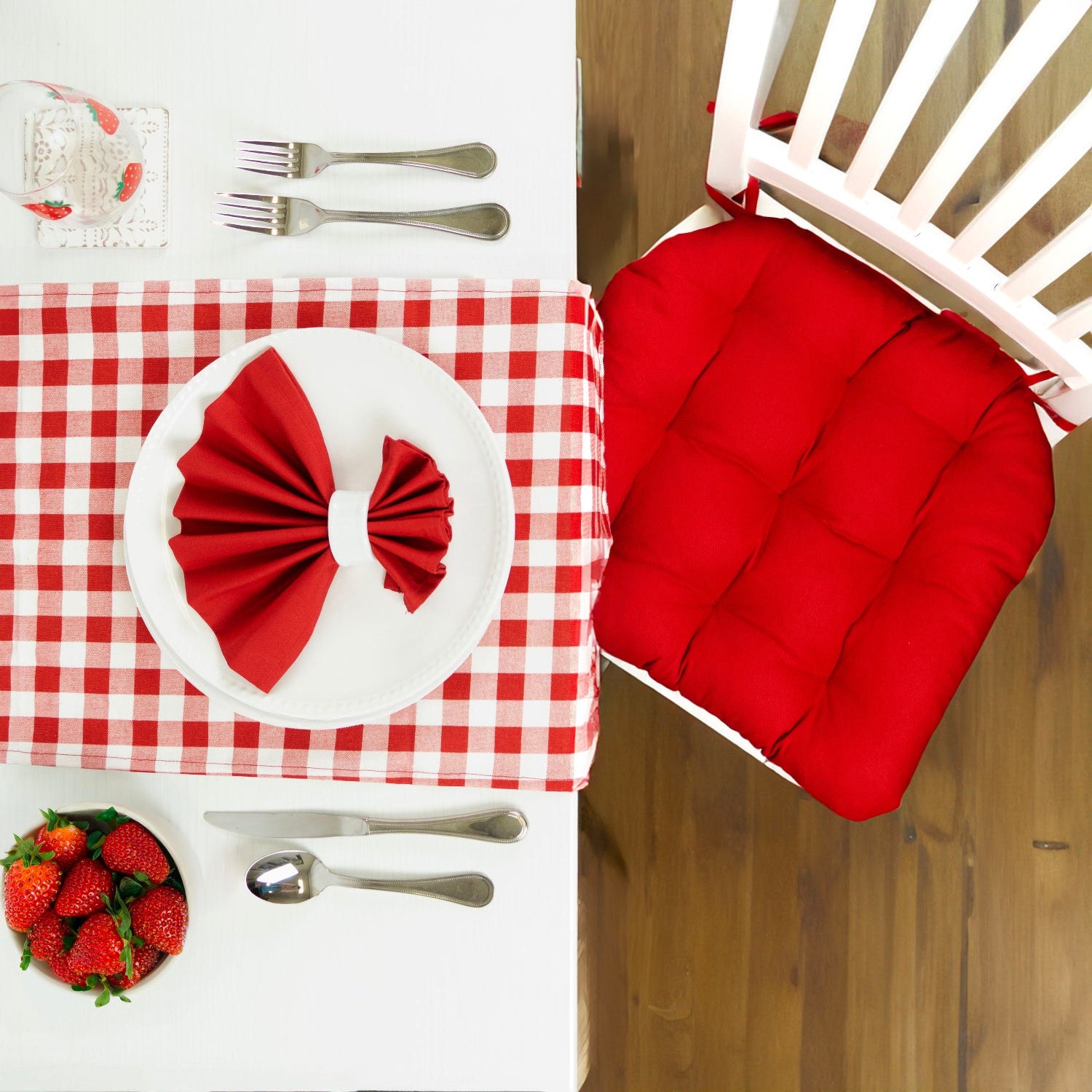 Red cushion on a chair next to a table with red and white checkered tablecloth, white plate with red napkin, and strawberries.